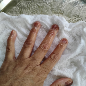 A close-up, overhead shot shows a person’s hand resting on a white, textured cloth, which is draped over the same ornate silver tray. The fingers and fingernails appear wet or glossy, as if treated with oil or water. The intricate floral etchings of the metallic tray are visible in the background, partially obscured by the thin, bunched fabric.